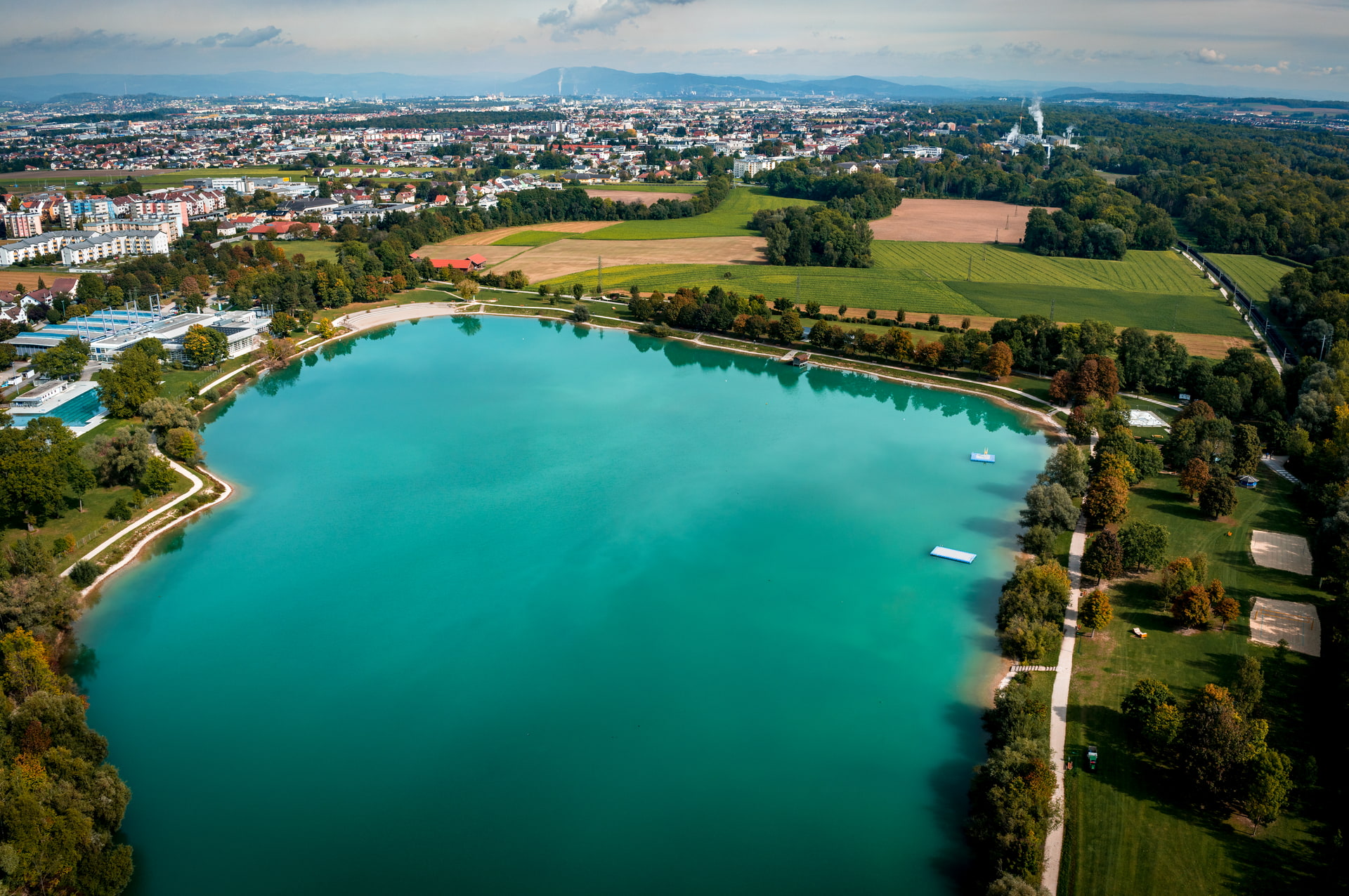 Luftaufnahme eines türkisfarbenen Sees mit Uferweg, umgeben von Feldern und einer Stadt im Hintergrund.