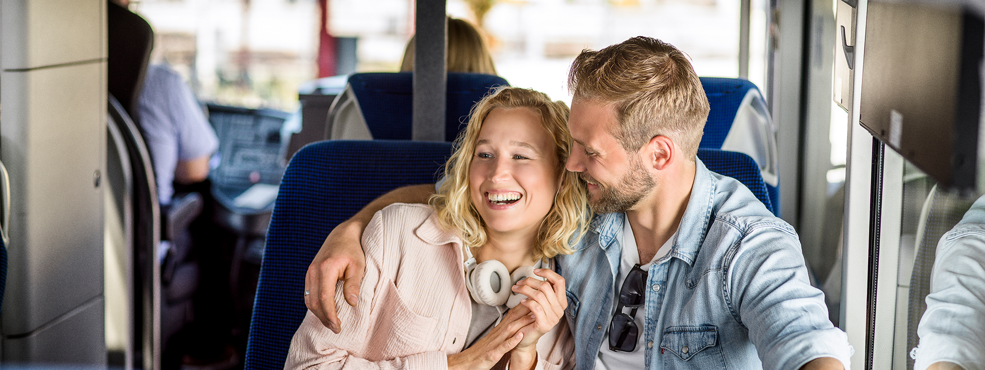 Paar sitzt eng beieinander im Bus, lacht, sie trägt Kopfhörer um den Hals.