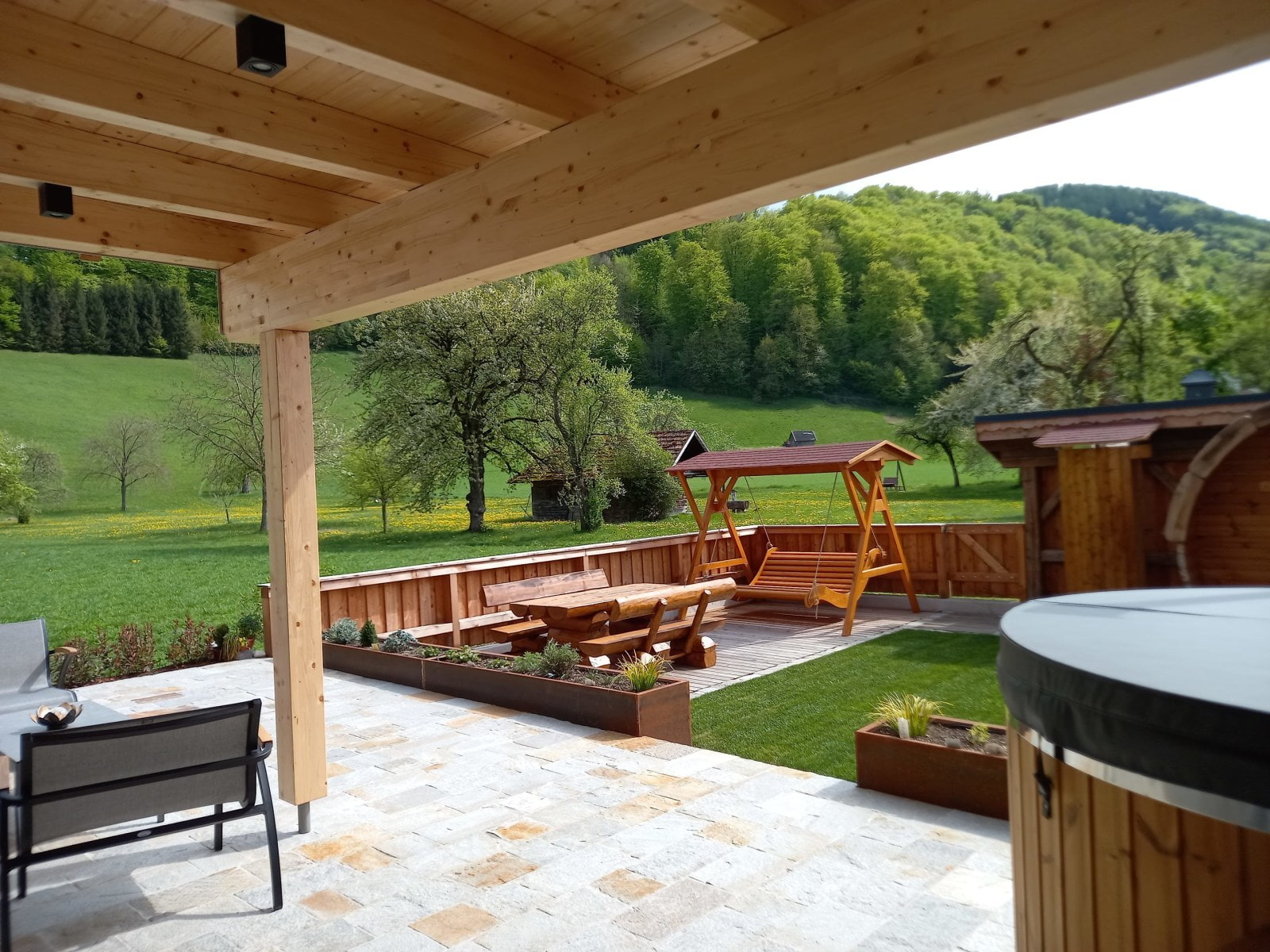 Eine Holzterrasse mit Sitzgruppe und Schaukel, daneben grüne Wiese, Bäume und sanfte Hügel im Hintergrund.
