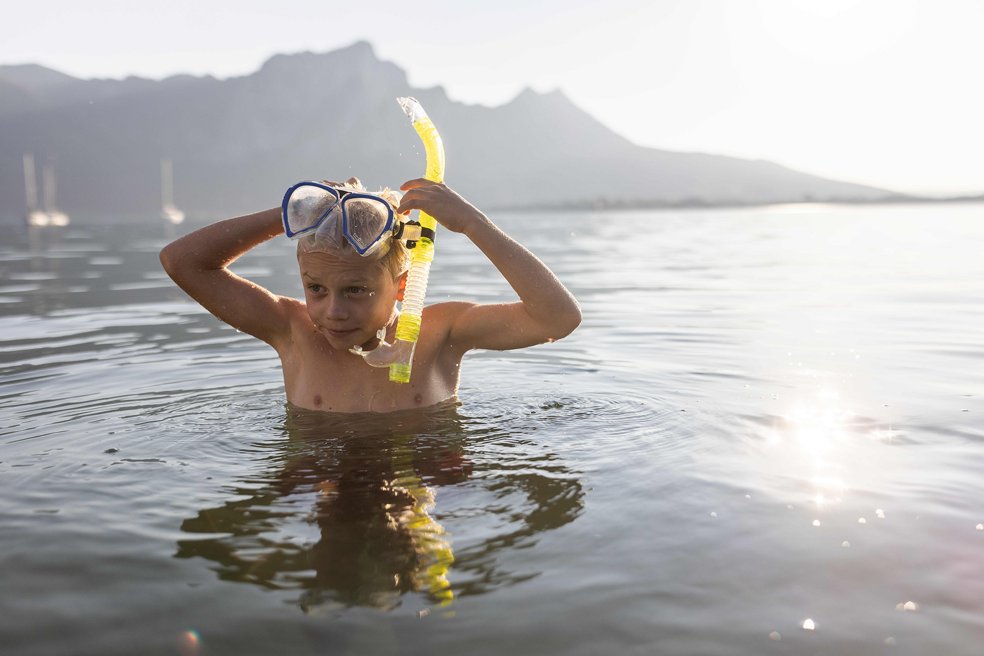 Junge beim Schnorcheln im Wasser, Maske und Schnorchel auf dem Kopf, Berglandschaft im Hintergrund.