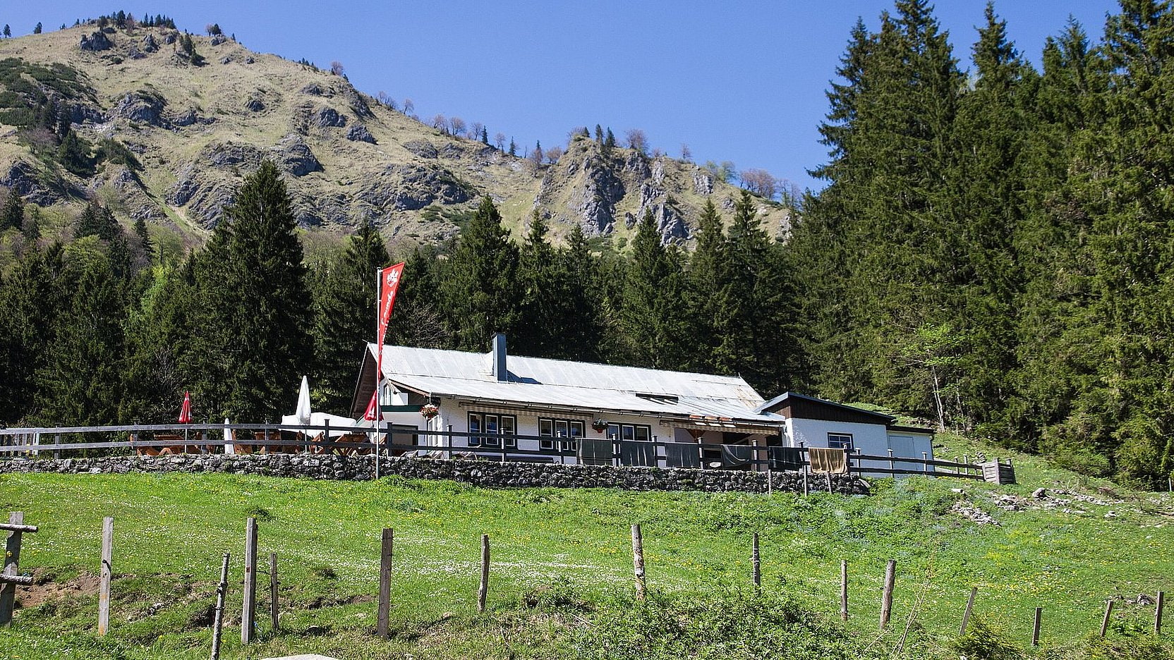 Berghütte im Grünen am Waldrand, vor felsigen Bergen unter blauem Himmel.