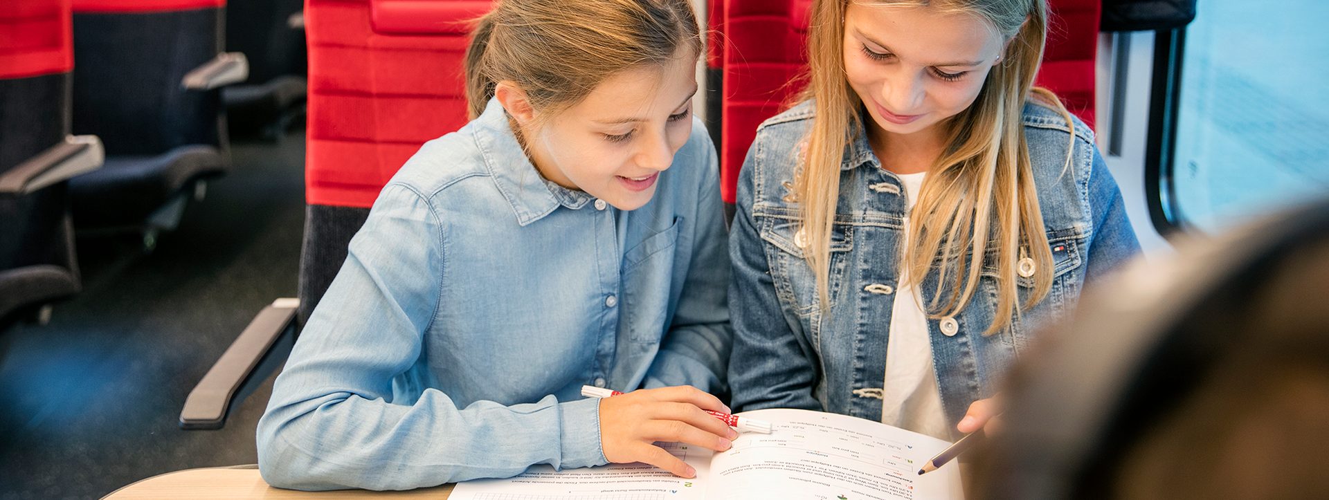 Two girls studying together at a table on a bus, looking at an open workbook.
