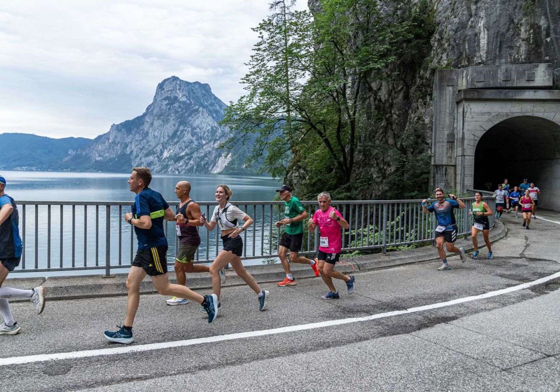 Läufergruppe joggt entlang der Seestraße mit Berglandschaft; rechts Felswand mit Tunnel.