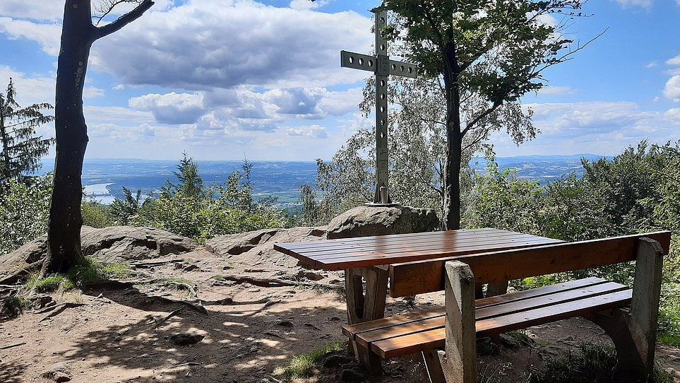 Ausflugstipp Puchenauer Kreuzweg Holztisch mit Bank auf felsigem Aussichtspunkt, Blick über Landschaft; Metallkreuz hinter dem Tisch, von Bäumen umgeben.