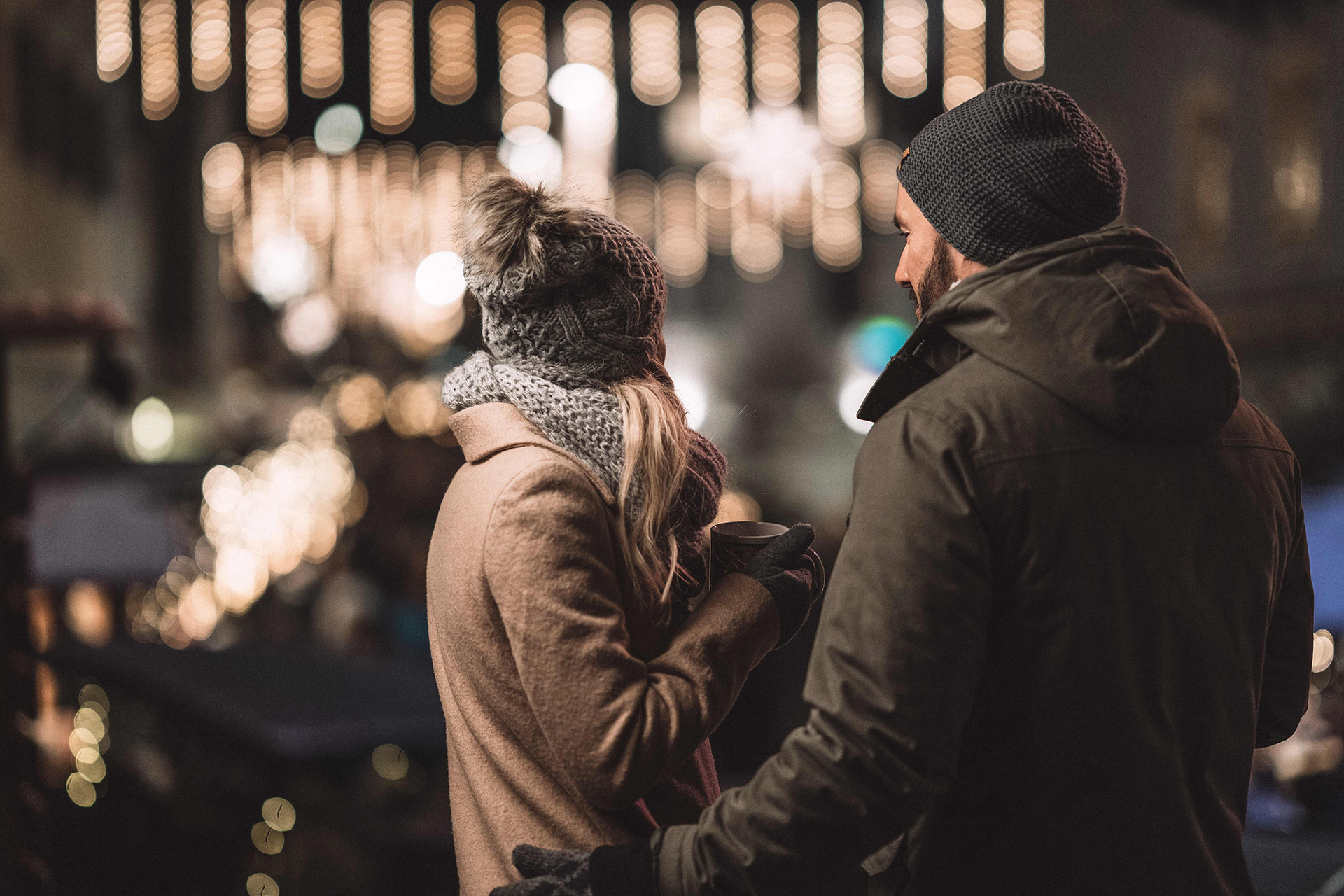 Ausflugstipp Adventmärkte Zwei Menschen im Winter stehen eng beieinander, halten warme Getränke; im Hintergrund funkeln Lichterketten.
