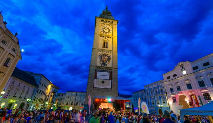 Ausflugstipp Enns Historischer Turm im Zentrum bei Nacht, Bühne und Menschenmenge auf dem Marktplatz.