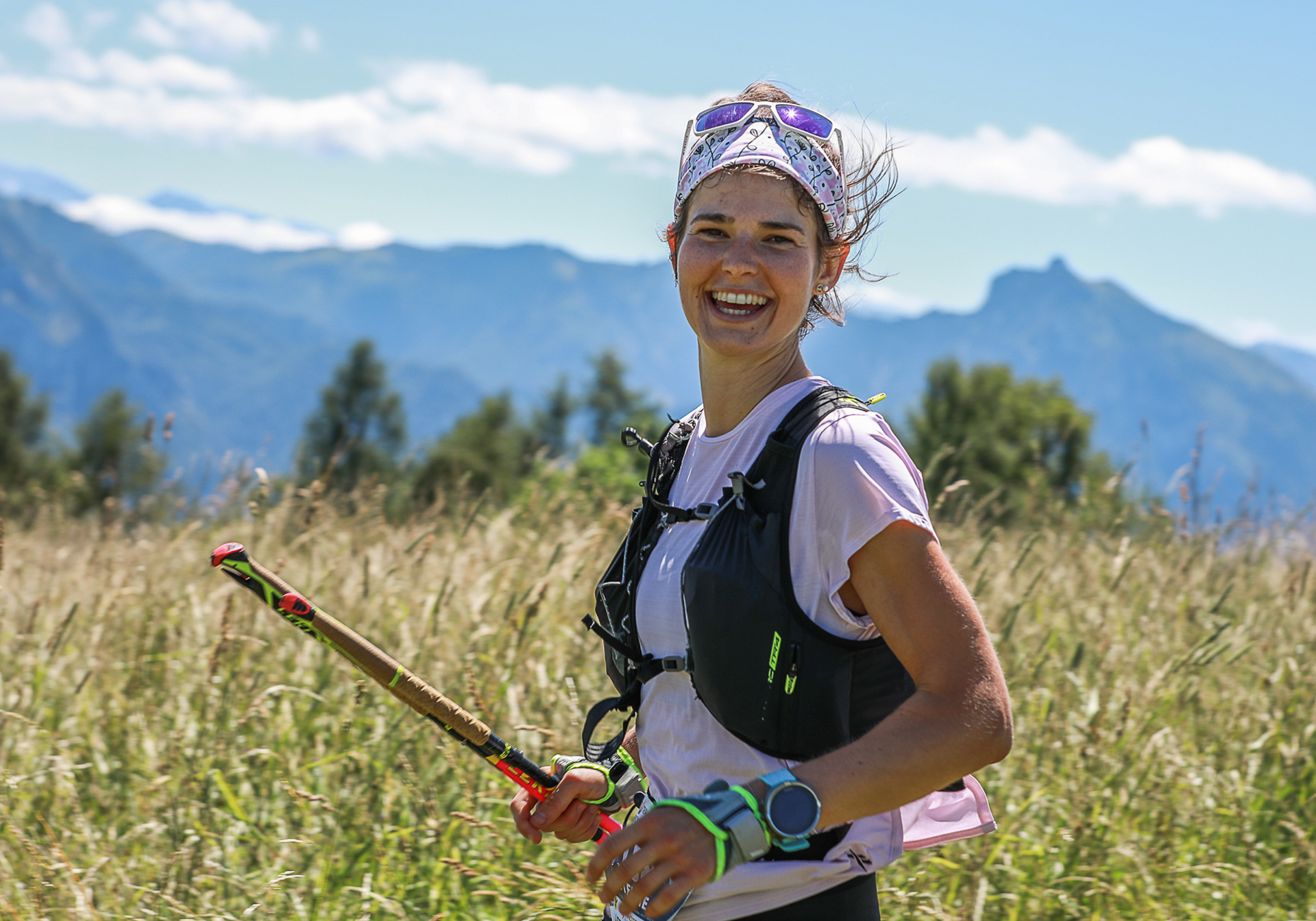 Smiling woman hiker with trekking poles in a grassy field, mountains in the background.