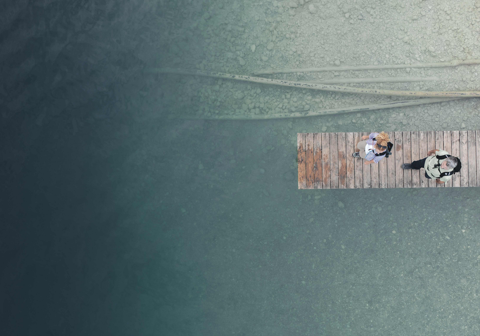 Zwei Personen stehen auf einem Holzsteg, der ins klare Wasser ragt.