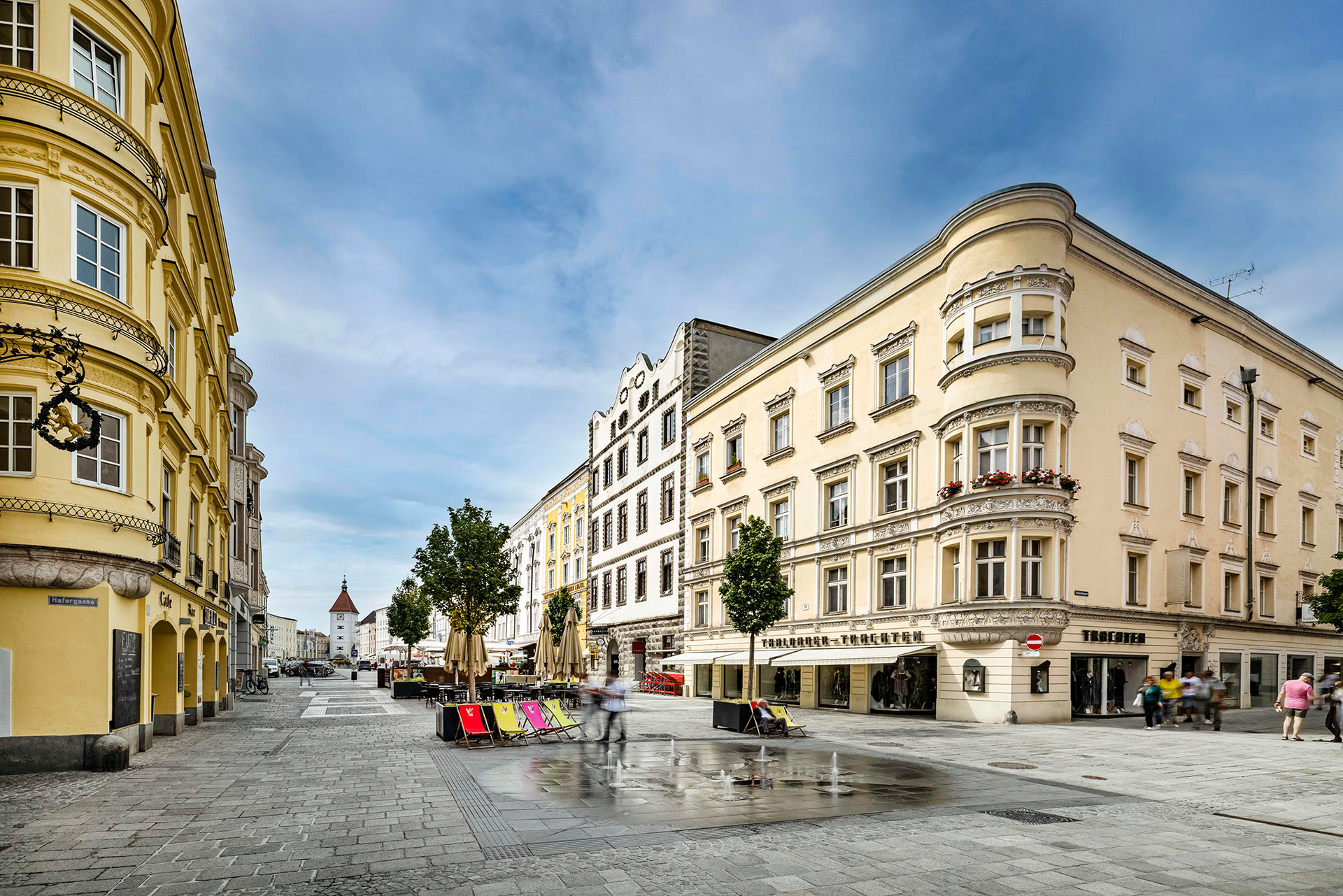 Historischer Stadtplatz mit pastellfarbenen Gebäuden, Straßencafés und Bäumen unter blauem Himmel.