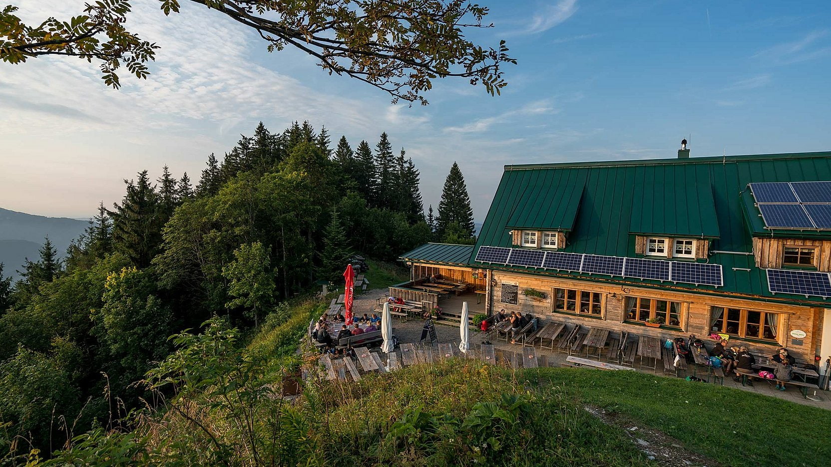 Ausflugstipp Ennserhütte Berghütte mit grünem Dach und Solarpanels, Terrasse voller Gäste, umgeben von Wald und Bergen.