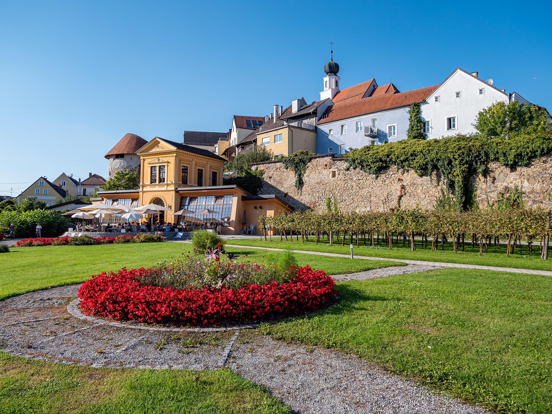 Historisches Gebäudeensemble mit gelbem Café, Terrassenbestuhlung, Blumengarten und Steinmauer.