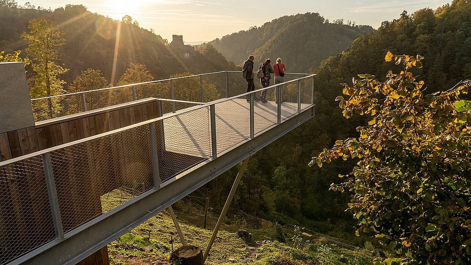 Ausflugstipp Jahresstiege Gramastetten Drei Personen stehen auf einer erhöhten Metall-Aussichtsplattform über einem bewaldeten Tal bei Sonnenuntergang.