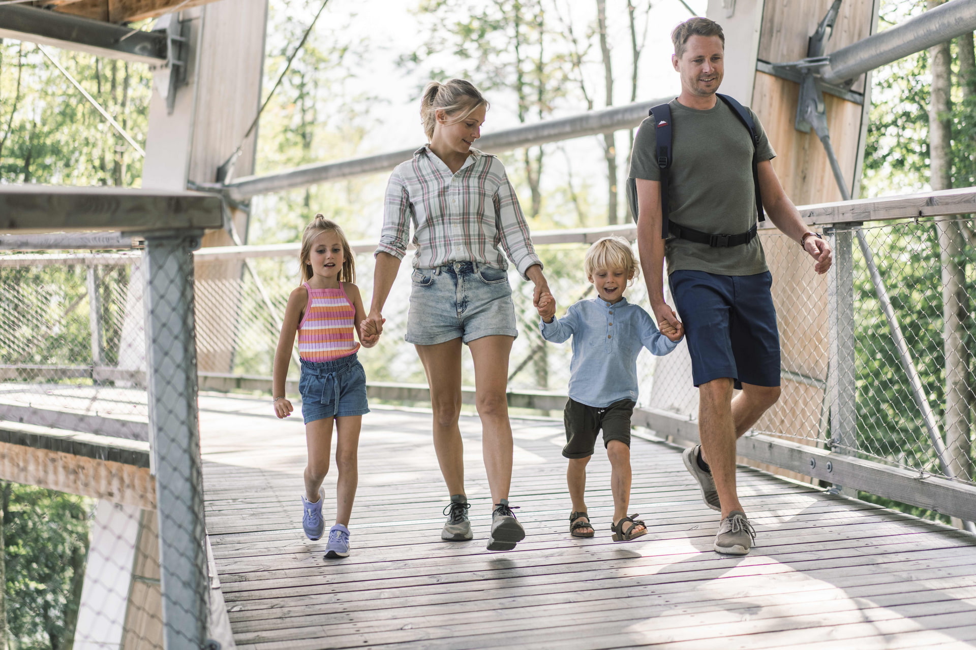 Baumwipfelpfad Gmunden Eltern mit zwei Kindern halten sich an den Händen und gehen über eine Holzbrücke im Park.
