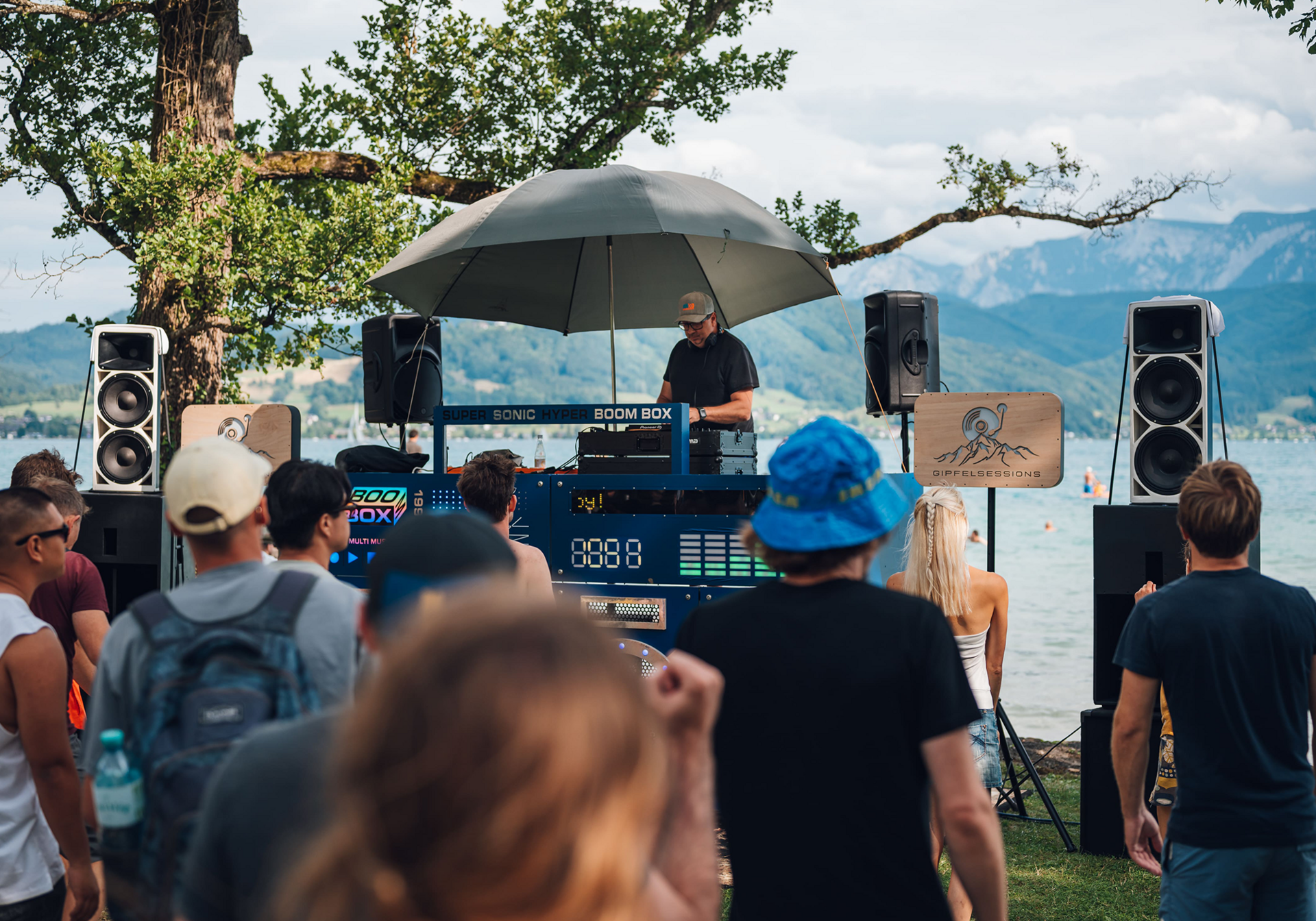DJ an einem Mischpult unter einem großen Sonnenschirm, umgeben von Lautsprechern, während Menschen am See zuhören.