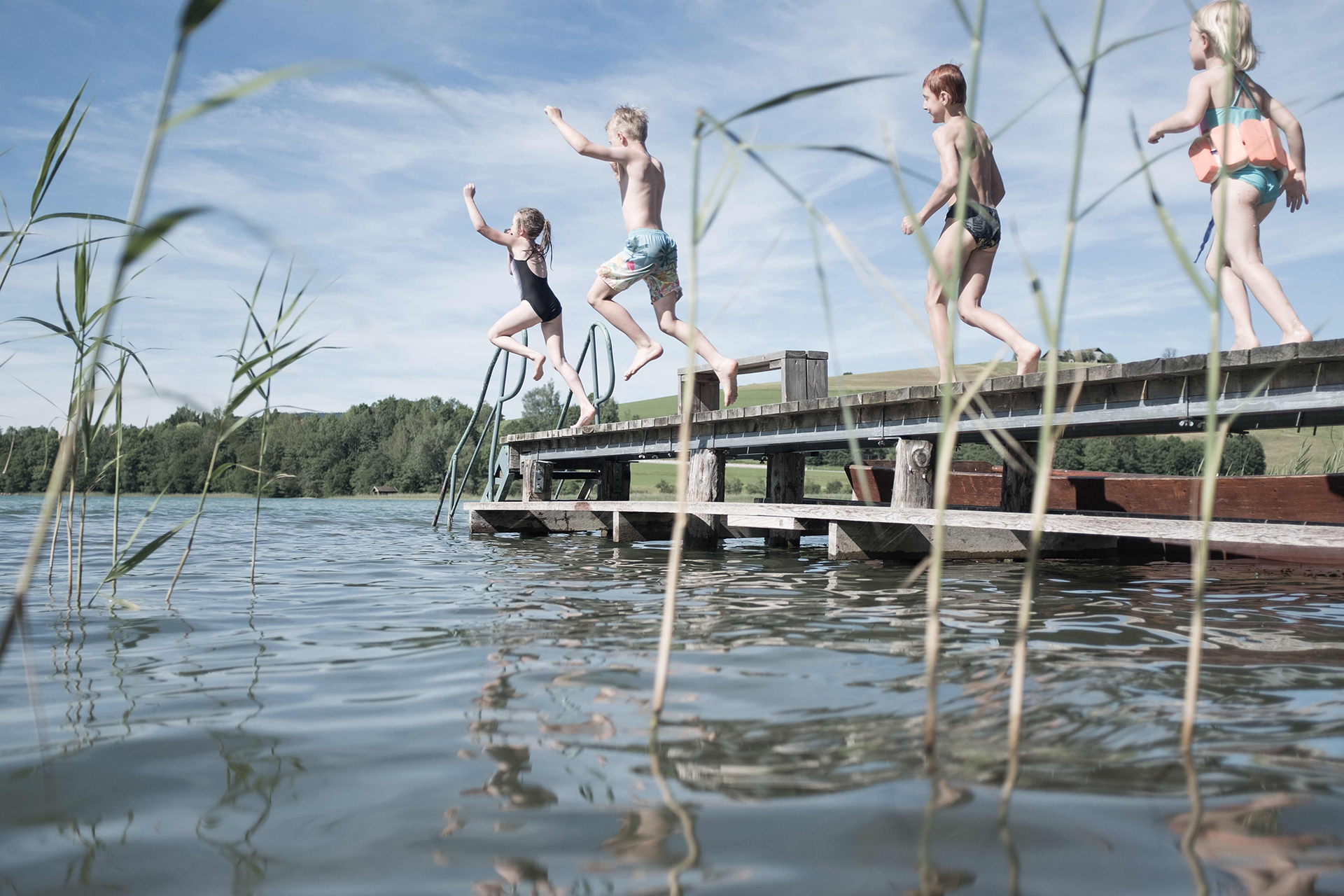 Mehrere Kinder springen vom Steg ins Wasser eines Sees bei sonnigem Himmel.