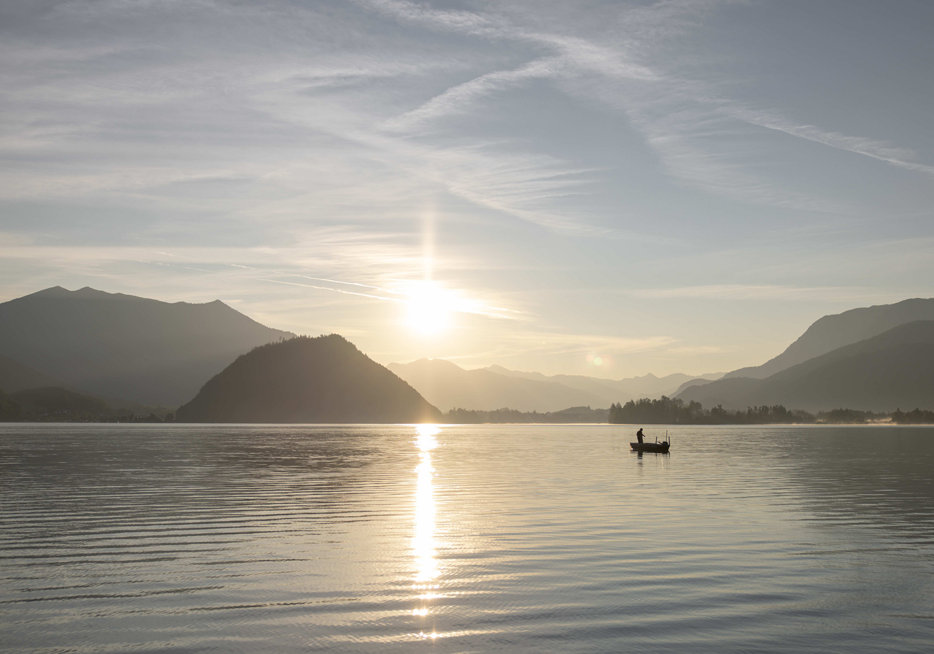 Ausflugstipp Wolfgangsee Landschaft eines Sees bei Sonnenaufgang mit Bergen im Hintergrund; eine Person fährt in einem kleinen Boot.