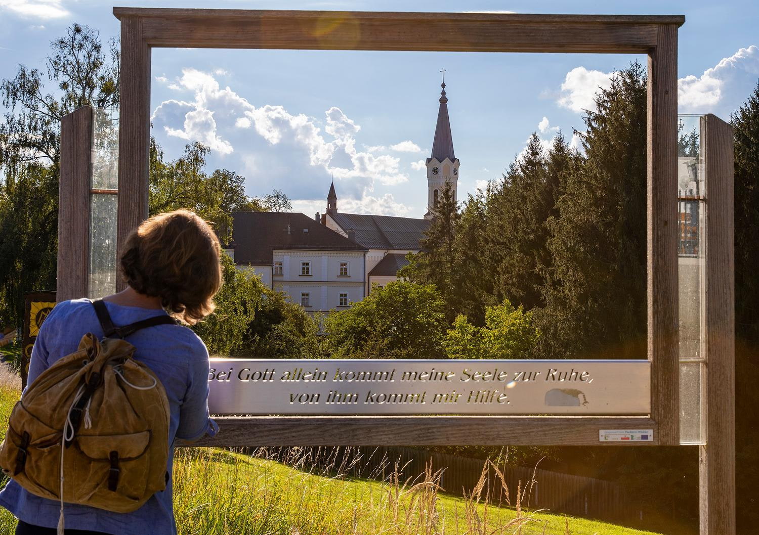 Ausflugstipp Maria Schmolln Landschaftsrahmenweg Quellenviertel Person mit Rucksack blickt durch einen Holzrahmen auf den Kirchturm hinter Baumlandschaft und Häusern.