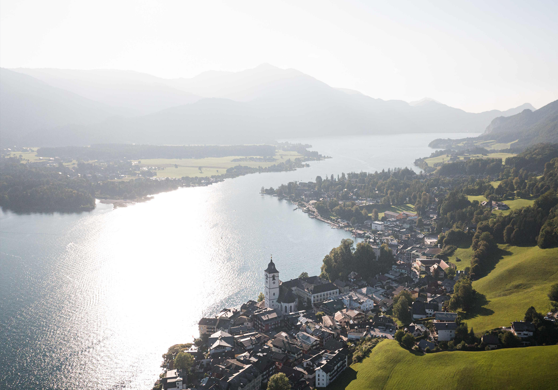 Luftbild eines Sees mit einer am Ufer liegenden Stadt und einem Kirchturm im Vordergrund vor Bergen.