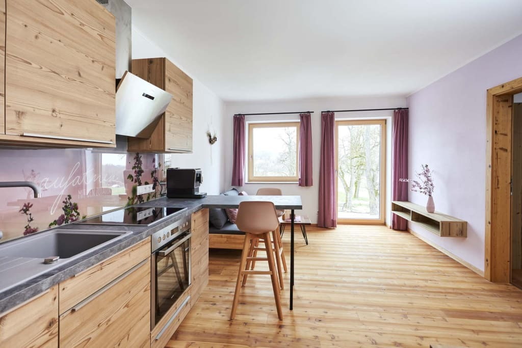 Mathiasnhof Offene Küche mit Holzmöbeln, Insel und Barhockern, Essbereich, große Fenster mit violetten Vorhängen und Holzboden.