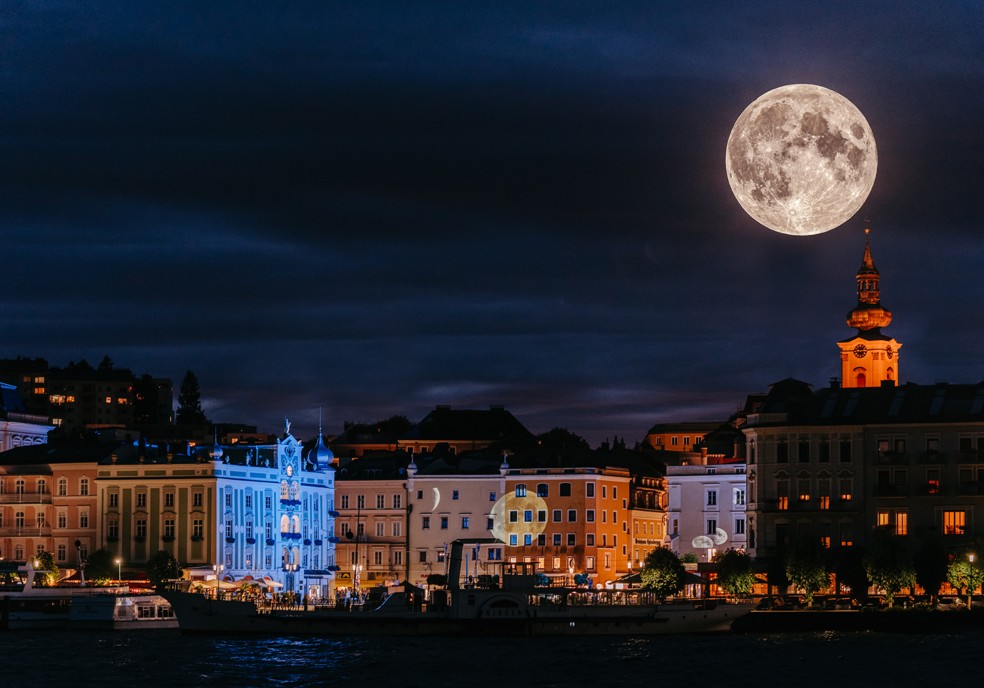 Großer Vollmond über einer Hafenstadt am nächtlichen Ufer mit beleuchteten Fassaden.