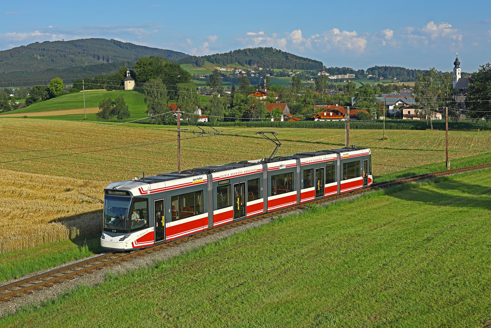 Moderne rote und weiße Straßenbahn fährt auf einer Bahnstrecke durch ländliche Feldlandschaft.