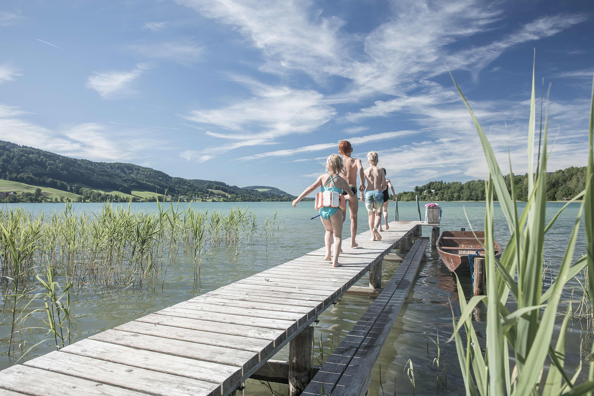 Familie geht barfuß über Holzsteg zum See; Kinder tragen Rettungswesten, Boot am Ufer.