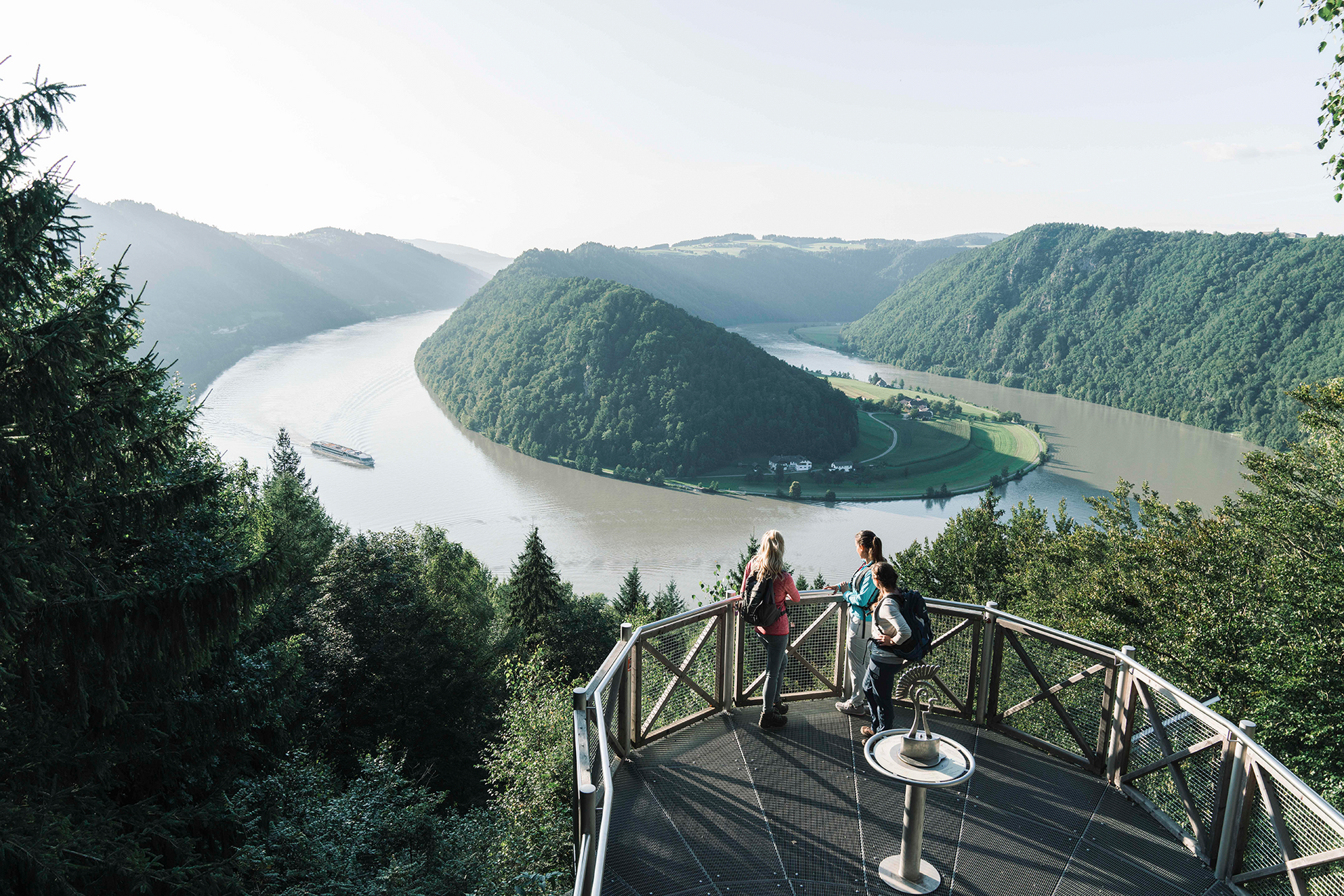 Ausflugstipp Donauregion OÖ Drei Personen stehen auf einer Aussichtsplattform und blicken auf den Fluss mit einer grünen Insel im Tal.
