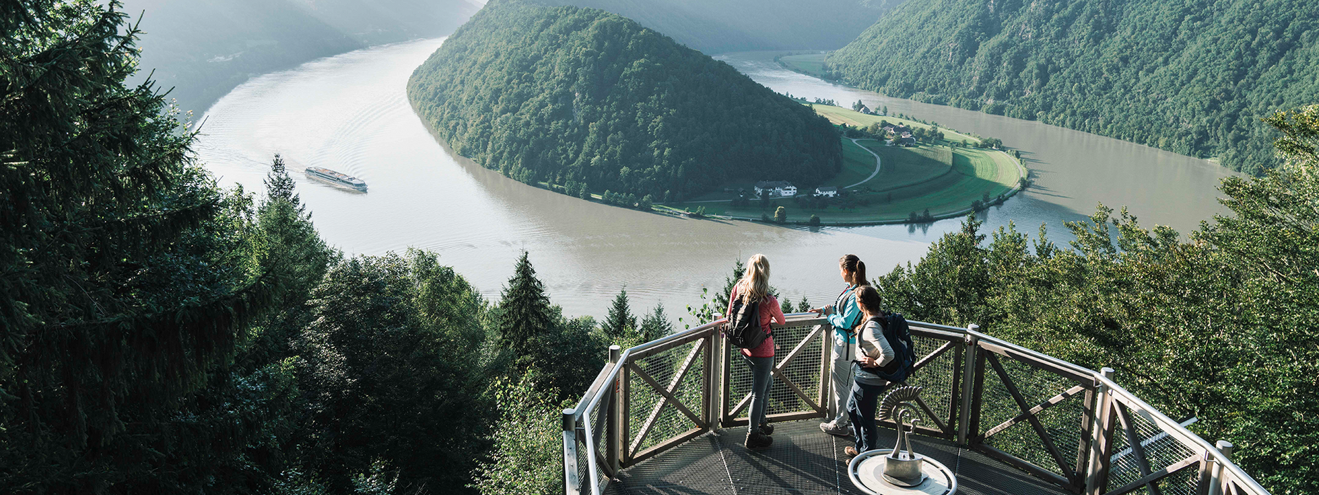 Drei Personen stehen auf einer Aussichtsplattform und blicken auf den Fluss mit einer grünen Insel im Tal.