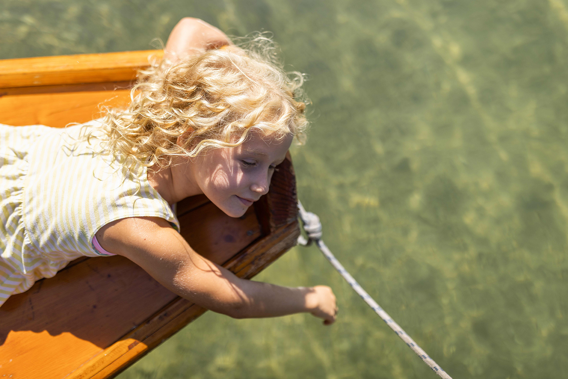 Junges Mädchen mit lockigem blondem Haar lehnt aus einem Holzboot und greift nach einem Seil.