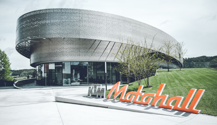 Ausflugstipp KTM Motohall Modernes, rundes Gebäude mit perforierter Metallfassade und Glasfront; orange Schriftzeichen davor auf grüner Wiese.