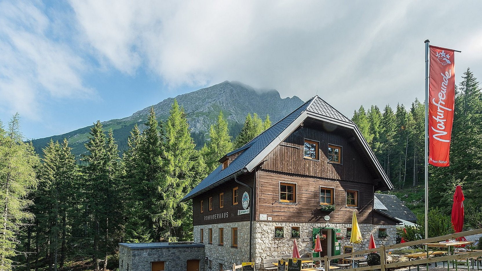 Alpine Berghütte aus Holz am Waldrand mit Bergen im Hintergrund.