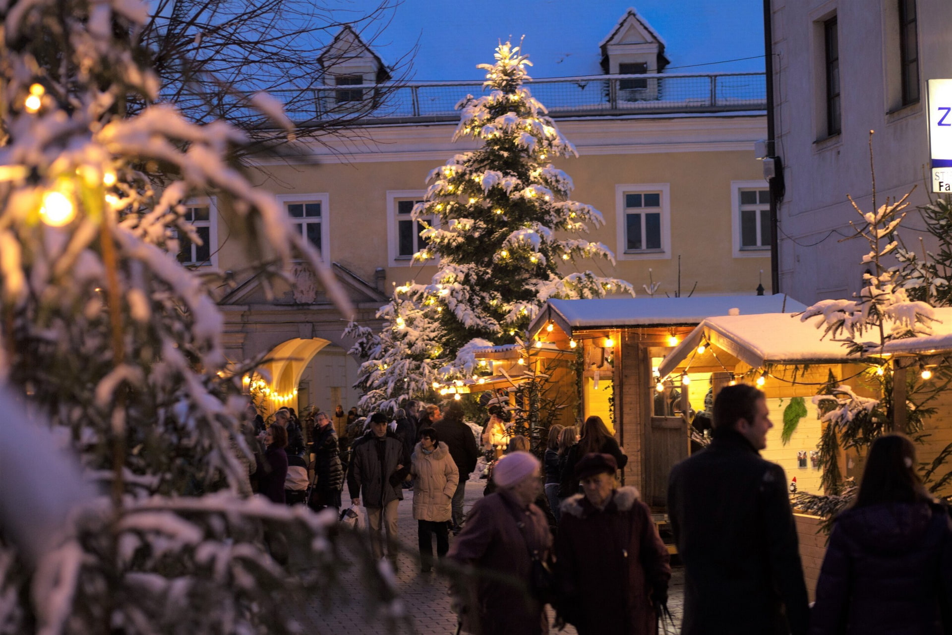 Weihnachtsmarkt im Abendlicht; Schnee bedeckt; leuchtender Baum; Holzhäuschen; Menschenmengen.