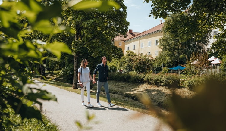 Paar geht Hand in Hand auf einem sonnigen Parkweg mit Bäumen und Häusern im Hintergrund.