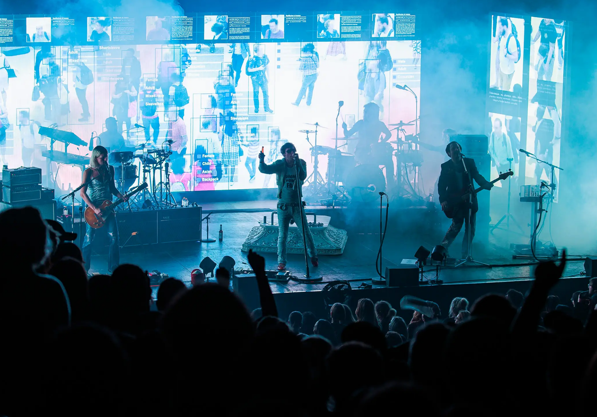 Band spielt auf Bühne; Leadsänger zentral, Gitarristen links/rechts; Nebel und LED-Bildschirme im Hintergrund.